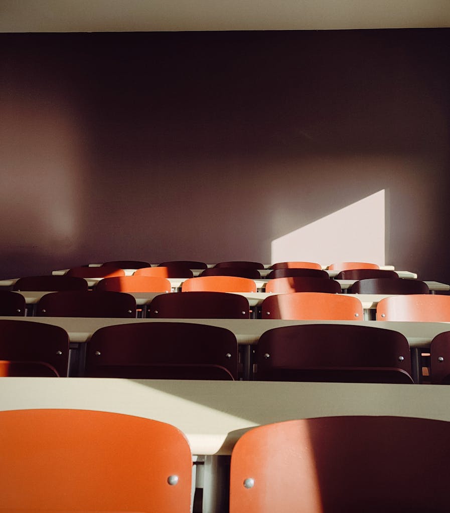 An empty classroom with sunlight casting shadows on red chairs, creating a dramatic effect.